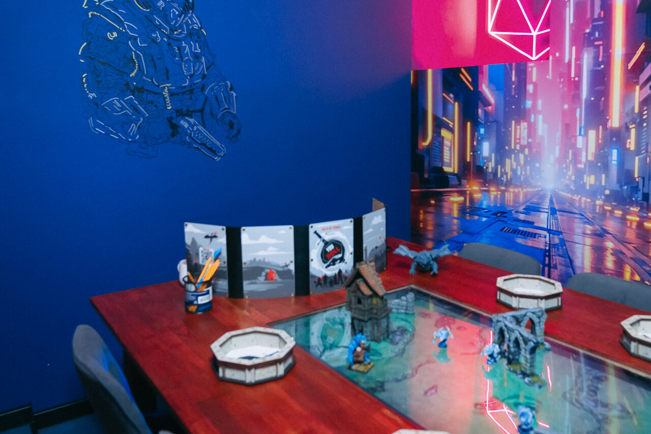 Арендовать YOU EVENT (Твой ивент). Клуб под закрытие. Комната для настольных игр с деревянным столом, на котором игровое поле и миниатюры. Синие стены, неоновая подсветка, фоновая картина с городским пейзажем. Несколько стульев вокруг стола.