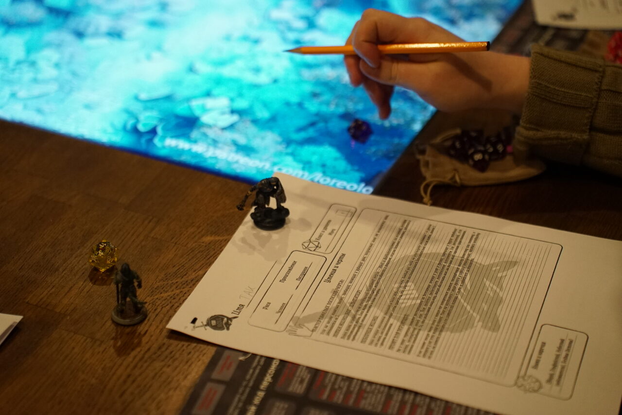Арендовать YOU EVENT (Твой ивент). Клуб под закрытие. Настольная игра на деревянном столе с листами бумаги, игральными фигурками и ручкой в руке человека. На заднем плане голубой светящийся экран. На листах бумаги виден текст и графика.