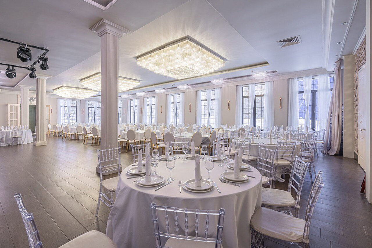 Арендовать Эдели. Просторный зал для мероприятий. Банкетный зал с круглыми столами, покрытыми белыми скатертями, прозрачными стульями с белыми подушками. Потолочные светильники с квадратными люстрами, деревянный пол, окна с белыми шторами, колонны, освещение дневное и искусственное.