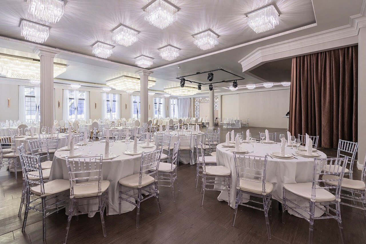 Арендовать Эдели. Просторный зал для мероприятий. Просторный зал с круглой расстановкой столов, покрытых белыми скатертями. Прозрачные стулья вокруг столов. Люстры подвешены к потолку, мягкое освещение. Светлые стены, темный пол. На дальнем плане - сцена с коричневыми занавесками.