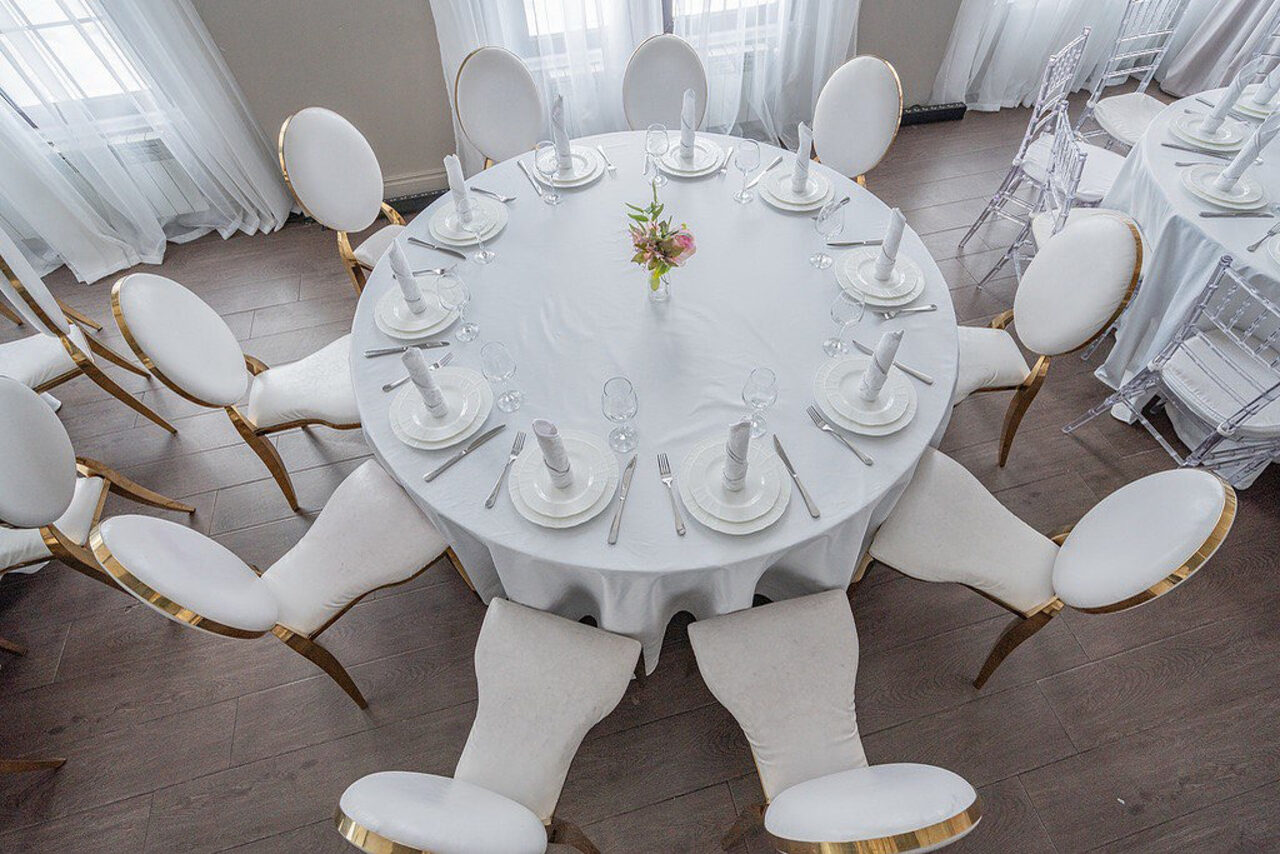 Арендовать Эдели. Просторный зал для мероприятий. Круглый банкетный стол с белой скатертью и сервировкой на 10 персон, включает тарелки, бокалы, столовые приборы и салфетки. В центре стола цветочная композиция. Вокруг стоят белые стулья. Пол деревянный, светлый. За окнами висят прозрачные шторы.