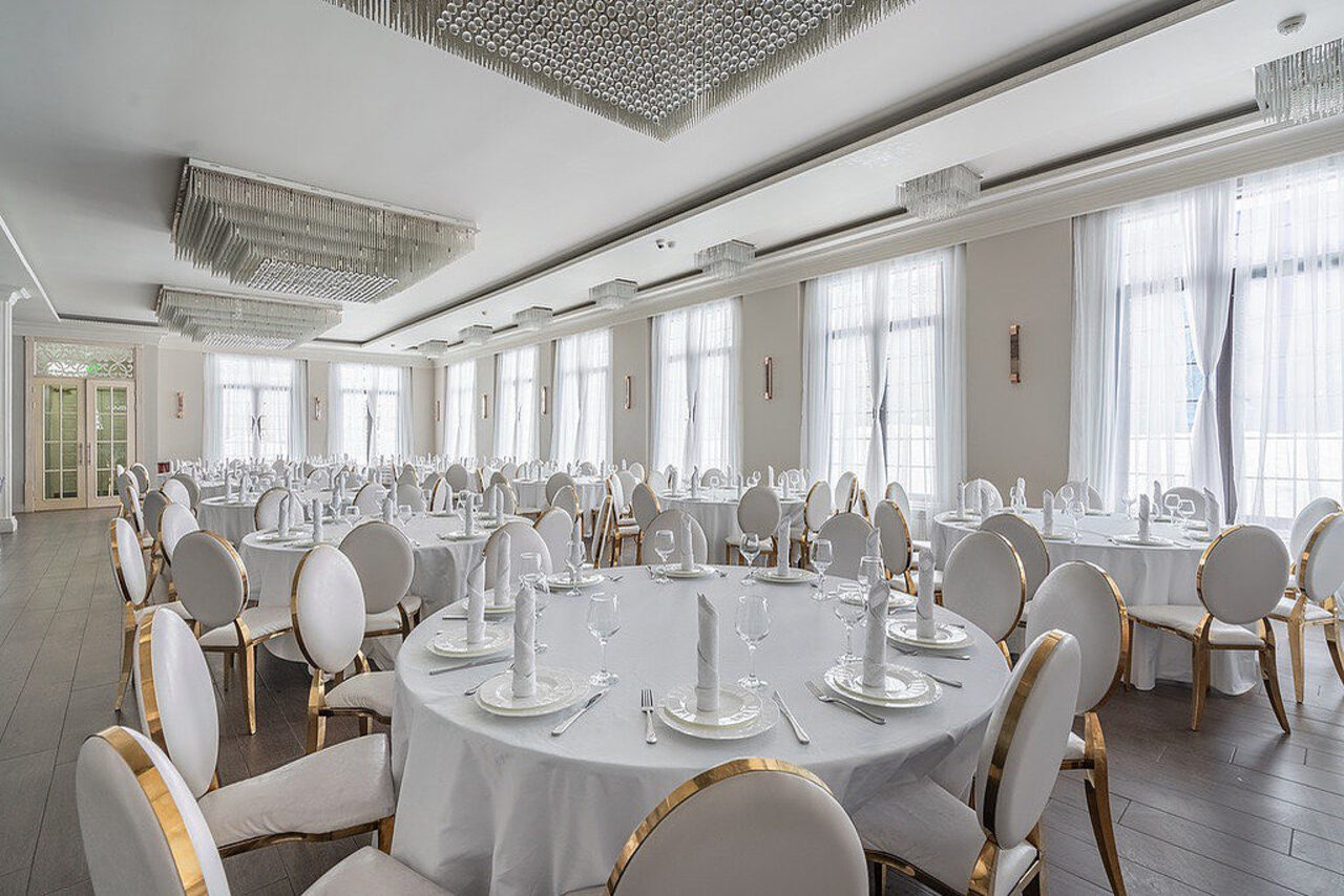 Арендовать Эдели. Просторный зал для мероприятий. Просторный банкетный зал с круглыми столами, белыми скатертями и стульями с мягкой спинкой золотистого цвета. Большие окна с белыми занавесками, потолочные светильники. Темный деревянный пол, белые стены. Столы сервированы приборами.