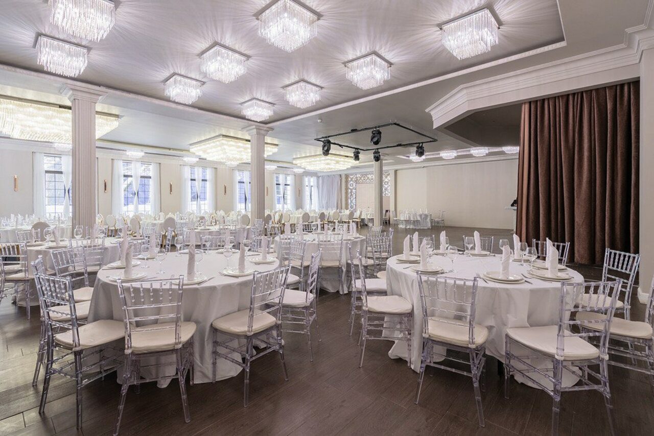 Арендовать Эдели. Просторный зал для мероприятий. Банкетный зал с белыми круглыми столами, прозрачными стульями, белыми скатертями и столовыми приборами. Большие окна, светлые стены, темный пол. Потолочные светильники с хрустальными элементами. Широкие колонны и коричневая штора.