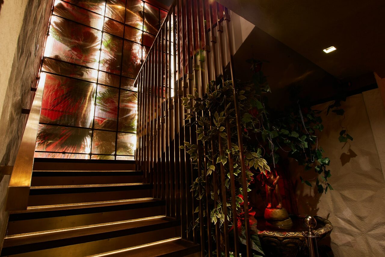 Арендовать Микс Afterparty(После вечеринки). Лестница с металлическими перилами, ведущая вверх. Витражное окно с абстрактным рисунком и подсветкой. Справа декоративное растение в вазе на столике. Стены светлые с текстурной отделкой. Нейтральное освещение.