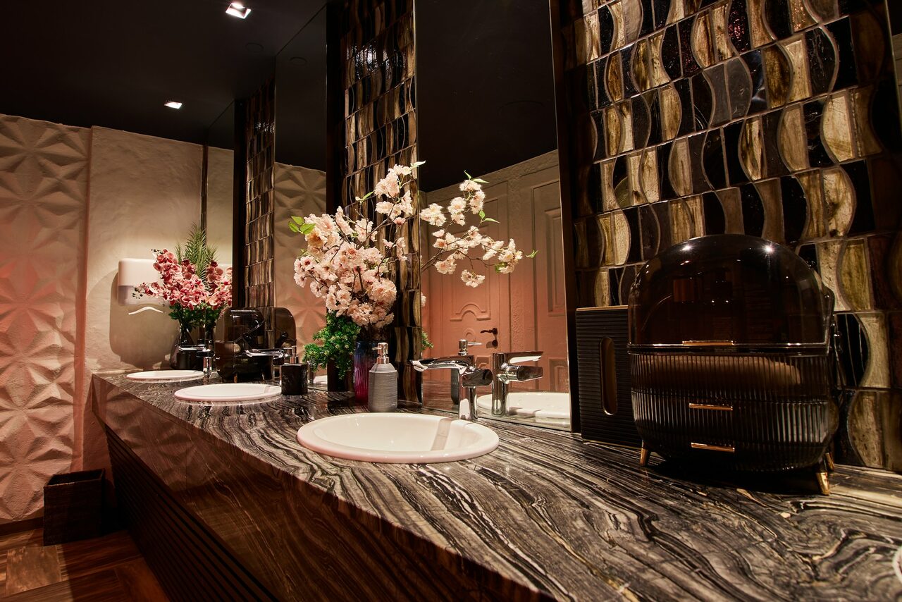 Арендовать Микс Afterparty(После вечеринки). Ванная комната с двумя раковинами, большие зеркала, столешница из темного мрамора с полосами, декоративные панели на стенах, флористические композиции в вазах, диспенсер для мыла, металлические смесители, бумажные полотенца, спокойное освещение.