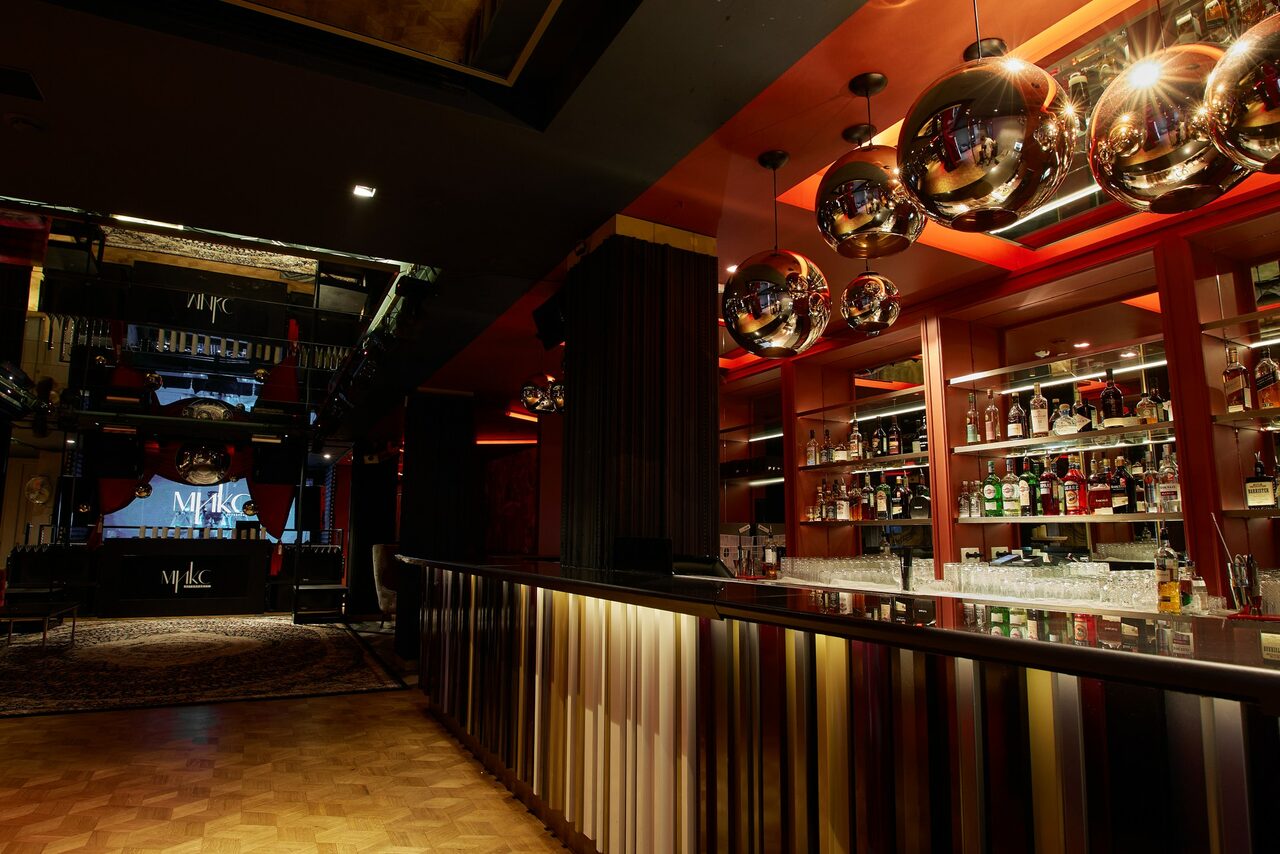 Арендовать Микс Afterparty(После вечеринки). Барное пространство с длинной стойкой, отделанной вертикальными полосами. На стене за стойкой полки с напитками. Потолок украшен круглыми металлическими подвесками. Пол деревянный, освещение теплое. Видно лестницу на второй этаж.