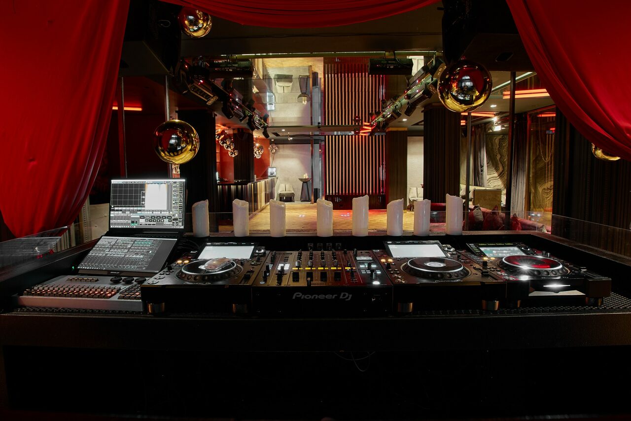 Арендовать Микс Afterparty(После вечеринки). DJ установка с микшером и двумя плеерами, окруженная красными занавесами и зеркальными шарами. Декорирование свечами, неоновое освещение и сочетание чёрного и красного цветов. Пространство с высокими стенами и глянцевыми поверхностями.