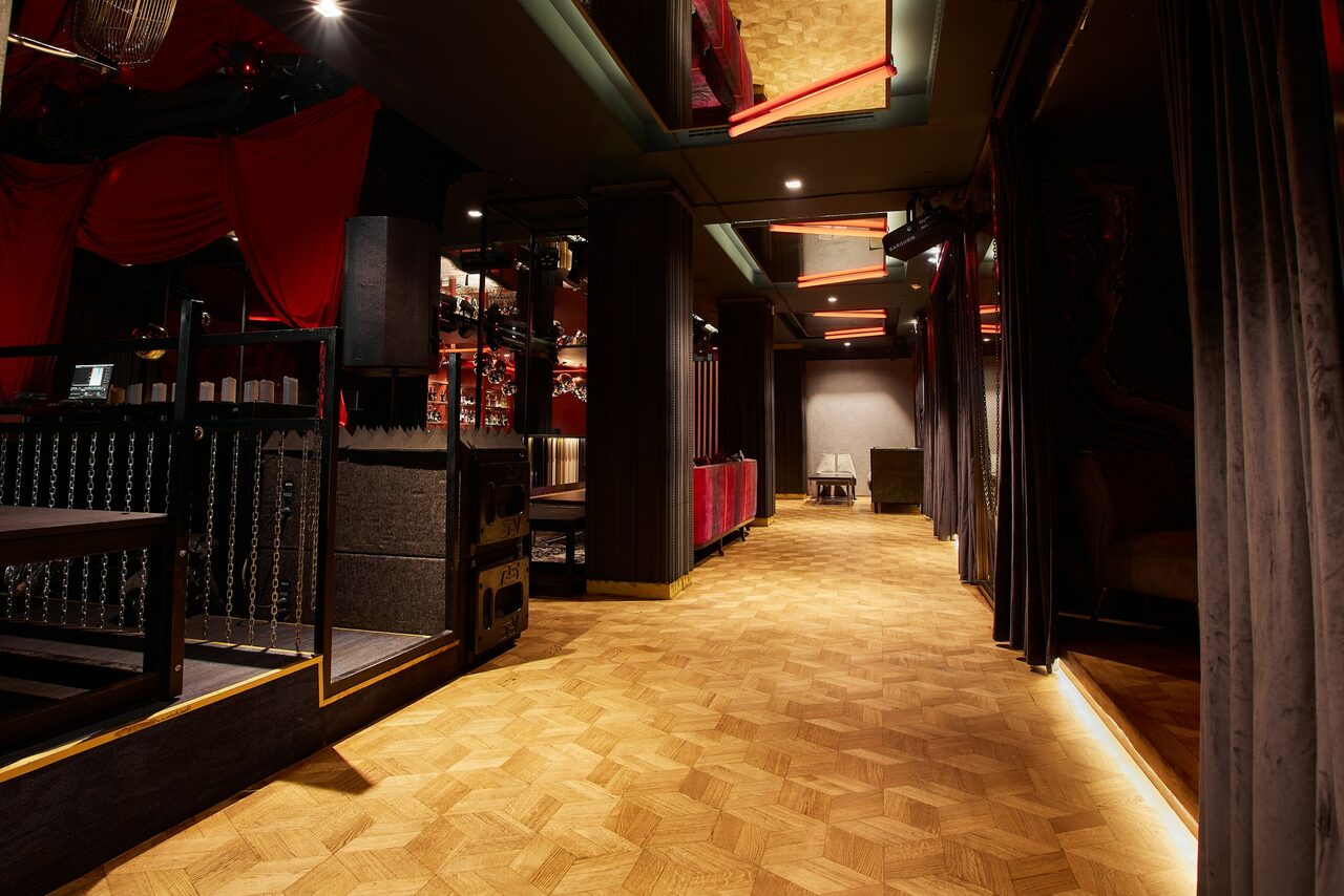 Арендовать Микс Afterparty(После вечеринки). Коридор с паркетным полом, справа драпировки серого цвета. Слева зона с черными и красными элементами, включая колонны и текстиль. Яркое точечное освещение, в конце коридора стол и стул у белой стены.