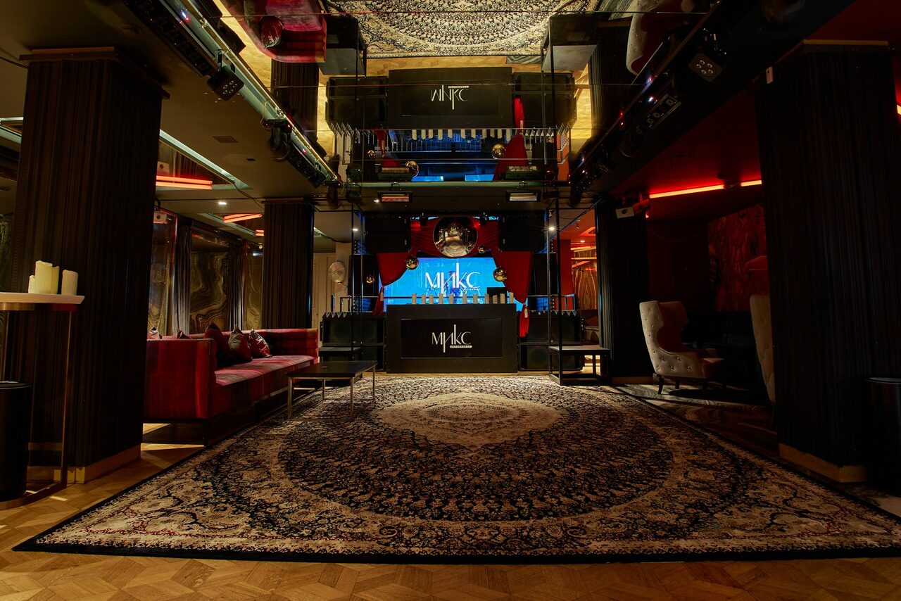 Арендовать Микс Afterparty(После вечеринки). Клубное пространство с деревянным полом. Большой восточный ковер. Красные диваны и кресла по периметру. Центральная стойка с логотипами и экранами. Пустяки и свечи на столах у колонн. Темное освещение с цветными акцентами. Потолочные прожекторы и зеркала.