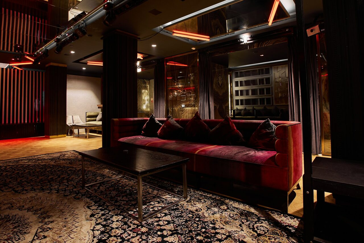 Арендовать Микс Afterparty(После вечеринки). Темное пространство с современным интерьером, большим красным диваном и черным ковром с узором. Стены с зеркальными и деревянными элементами. Красные и белые акценты в освещении. Чёрные и стеклянные столики. Потолок с неоновым декором.
