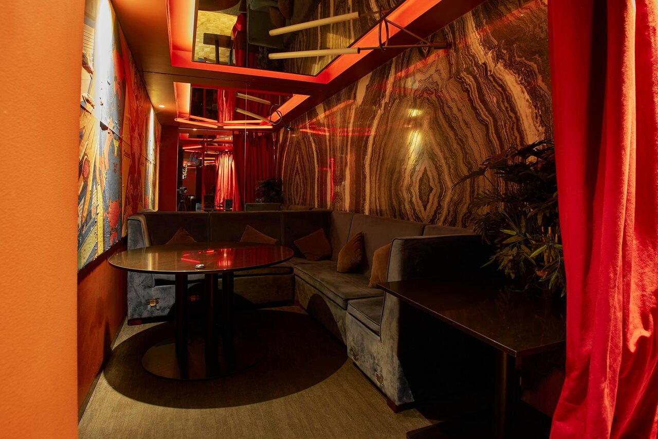 Арендовать Микс Afterparty(После вечеринки). Узкое помещение с оранжевыми и красными стенами, зеркальным потолком. Угловой диван серого цвета с подушками, круглый стол. Яркое красное освещение, стены с узором под мрамор, красные шторы. Декоративное растение в углу.