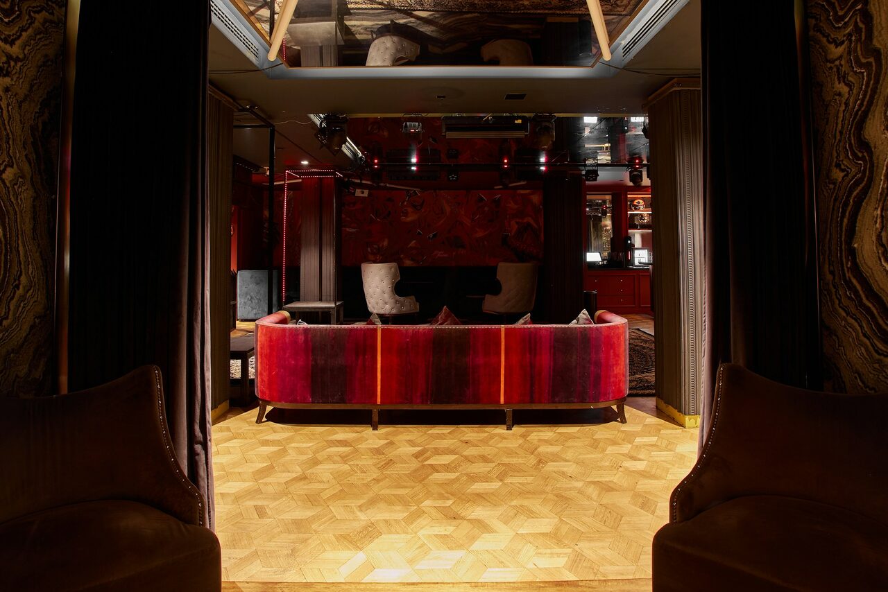 Арендовать Микс Afterparty(После вечеринки). Уютное пространство с паркетным полом, красно-бордовыми диванами, стульями и креслами. Мягкое освещение, тёмные стены с декоративными элементами. Зеркало на потолке, открытый бар на заднем плане, полки с бутылками.