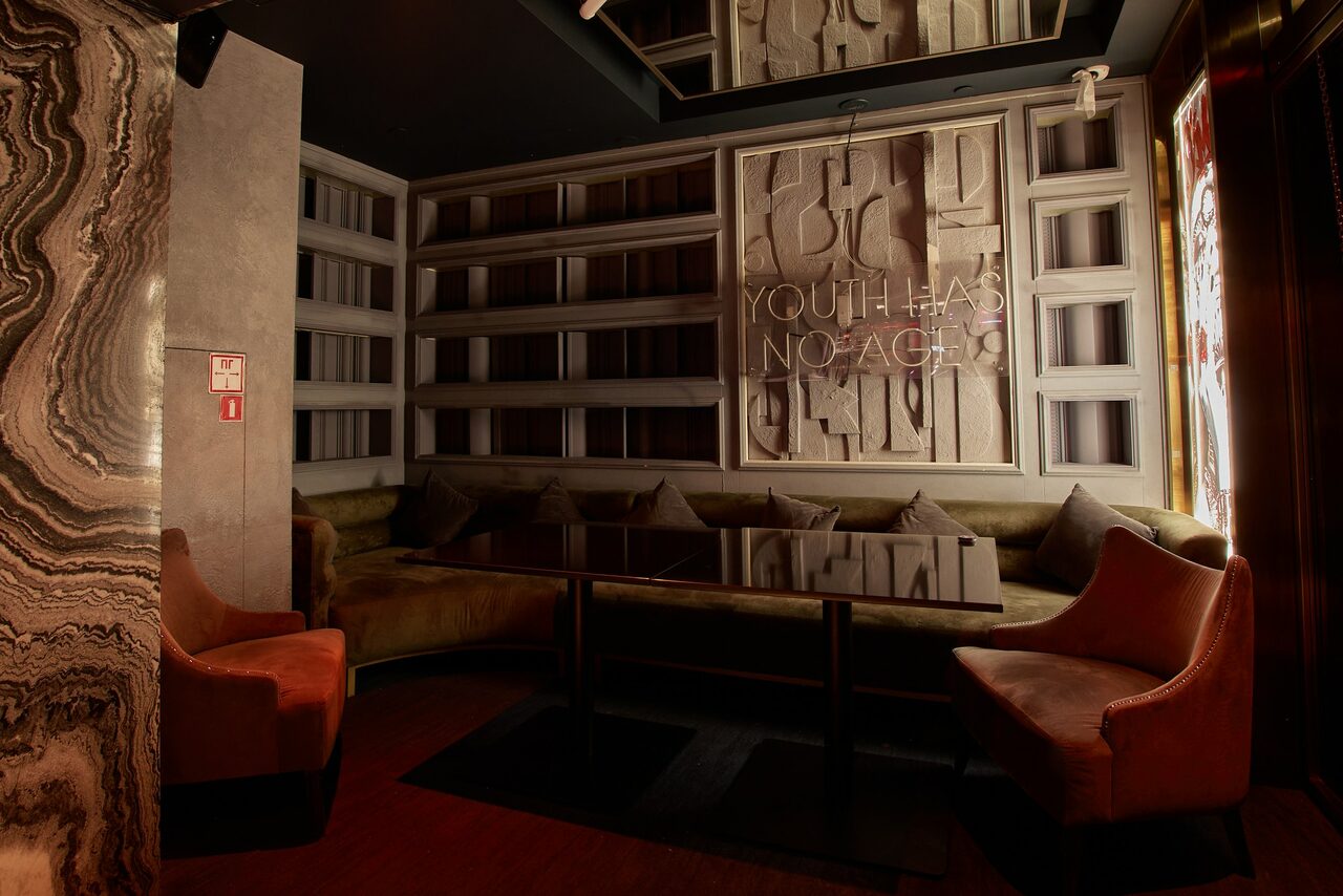 Арендовать Микс Afterparty(После вечеринки). Полуоткрытое пространство с мягкими диванами и креслами. Темный деревянный пол, стены с панелями и встроенными полками. Центральный стеклянный стол. На стене надпись "Youth Has No Age". Неяркое освещение. Стена с мраморным рисунком.
