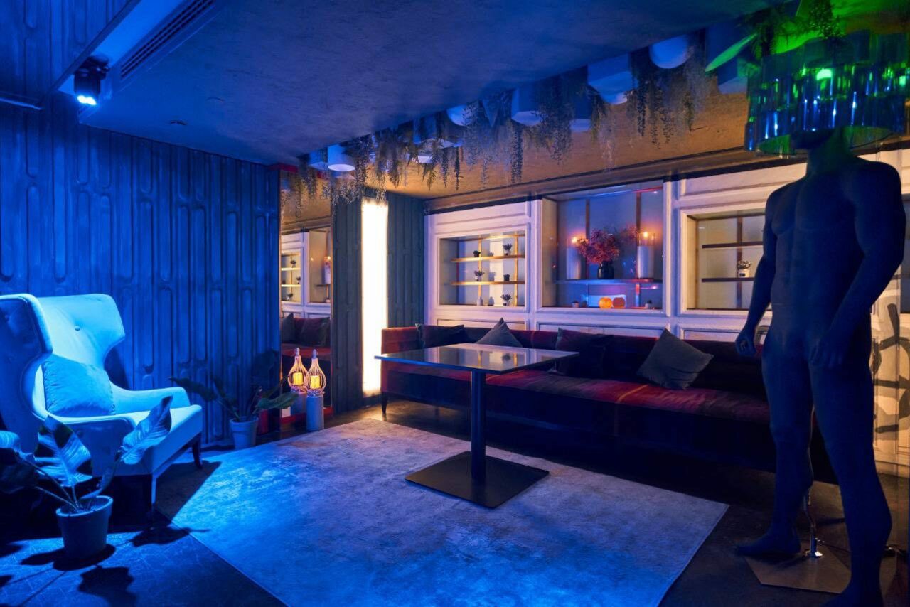 Арендовать Микс Afterparty(После вечеринки). Лофт-пространство с синим освещением, белое кресло, черный стол, темный угловой диван с подушками, большой ковёр, манекен, полки с декором, лампы, растения, деревянные панели и декоративные свисающие элементы на потолке.