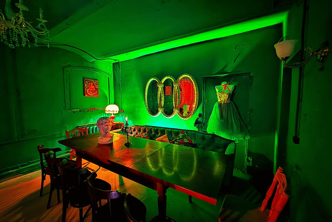 Арендовать Лофт «Рич» (RICH). Комната в зеленых тонах с длинным деревянным столом и стульями. На стене три овальных зеркала, манекен с платьем, картина. На столе настольная лампа и ваза. Потолочный светильник и бра на стене. Пол светлого дерева, стены и потолок зеленые.