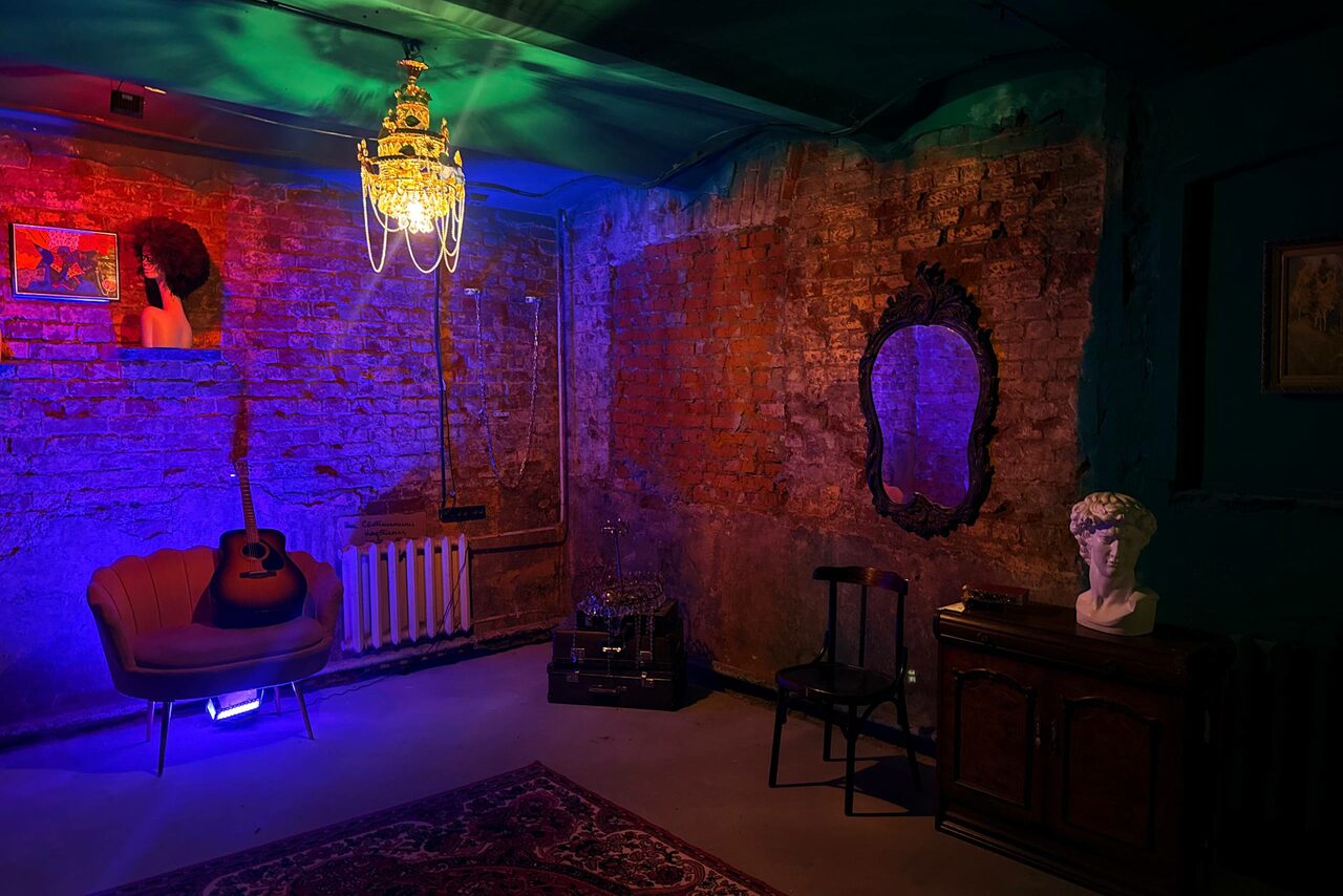 Арендовать Лофт «Рич» (RICH). Подвальное помещение с кирпичными стенами, креслом, гитарой и бюстом на комоде. Винтажное зеркало, деревянный стул и ковёр. Потолочный светильник. Освещение разноцветное, приглушенное. На стенах картины.