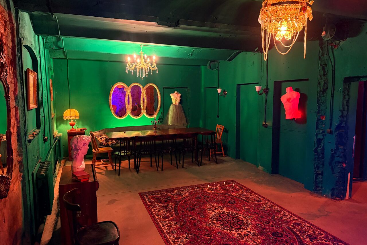 Арендовать Лофт «Рич» (RICH). Комната с зелёными стенами и потолком, длинный деревянный стол с разными стульями, на манекене платье, несколько бюстов, декоративные зеркала, люстра и настольная лампа. Пол покрыт красным ковром с узором. Освещение теплое и разноцветное.
