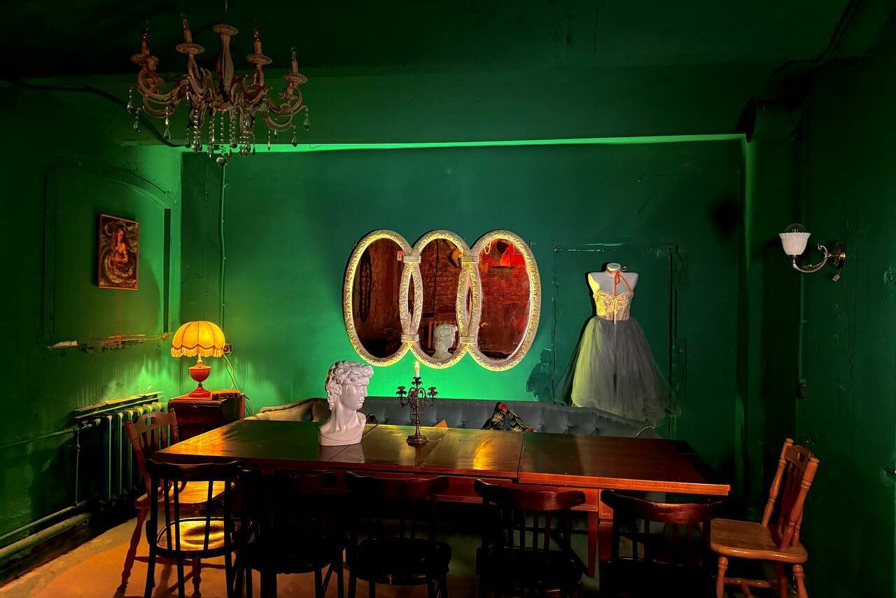 Арендовать Лофт «Рич» (RICH). Затемненное помещение с зелеными стенами. Большой деревянный стол окружен стульями. На столе бюст. На стене овальные зеркала и картина. В углу манекен с платьем. Люстра и настольная лампа обеспечивают освещение.