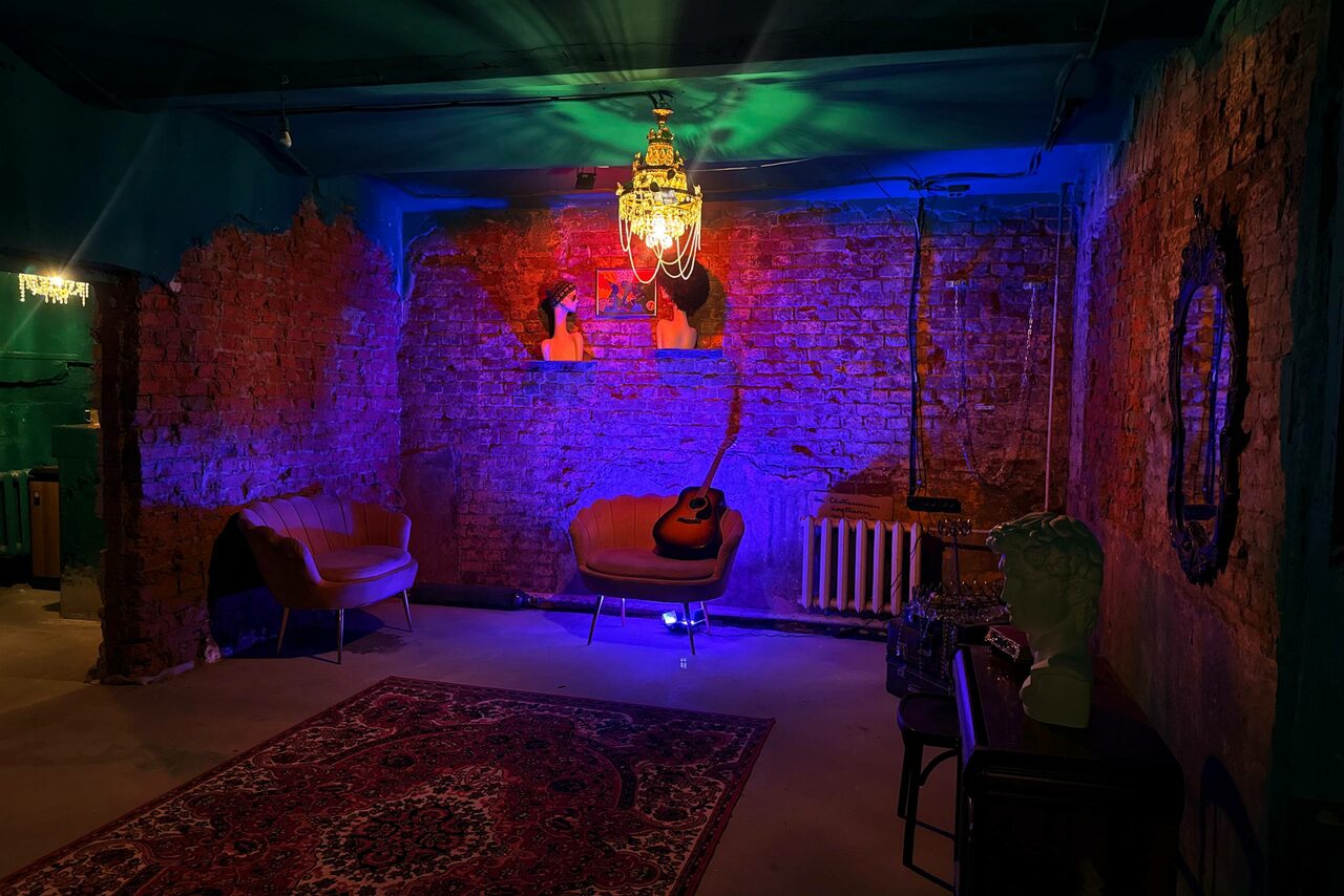 Арендовать Лофт «Рич» (RICH). Полуподвальное помещение с открытой кирпичной кладкой, двумя креслами, гитарой, декоративной люстрой и настольной лампой. На полу ковер, на стене зеркало. Освещение разноцветное, с акцентом на синий и зелёный. Потолок и стены зелёного цвета.