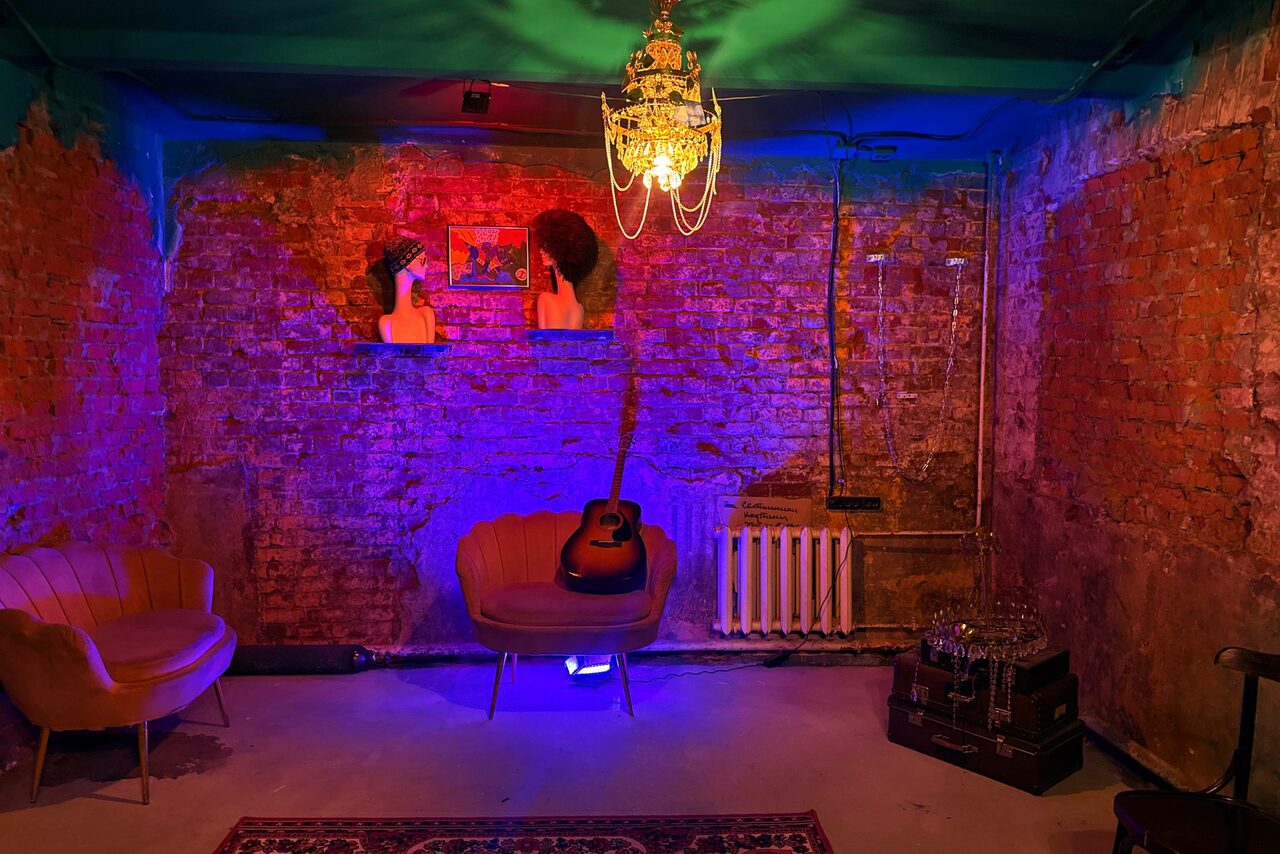 Арендовать Лофт «Рич» (RICH). Полуразрушенное кирпичное пространство с красноватым освещением и люстрой. Две фигурные статуи на полке, кресло, гитара на стуле, старый радиатор, деревянный ящик, трубы. Потолок темно-зеленый. Общий небрежный индустриальный стиль.