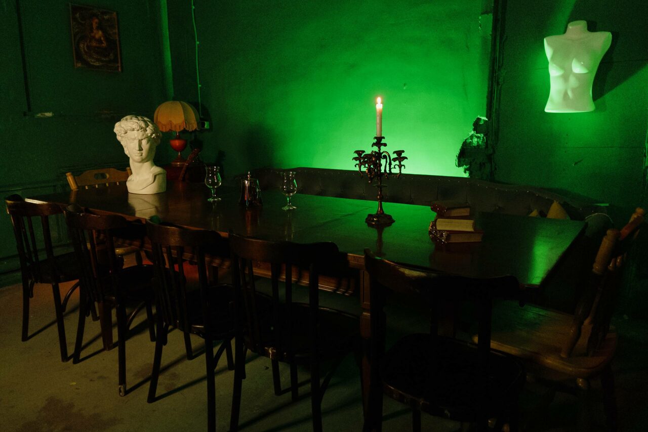 Арендовать Лофт «Рич» (RICH). Длинный деревянный стол с несколькими стульями, бюстом и канделябром. Стены и потолок зелёного цвета, слабое освещение. На стене висит торс, светящийся зелёным. На столе книги и бокалы. Слева торшер. Пол бетонный.