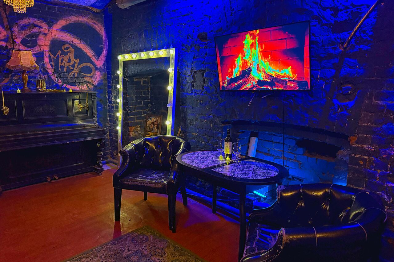 Арендовать Лофт «Рич» (RICH). Темное помещение с кирпичными стенами, подсвеченными синим светом. Две кожаные кресла, небольшой стол, игрушечный камин. На стене экран с изображением огня, зеркало с лампочками. На столе бутылка и бокал. Стены украшены рисунками.