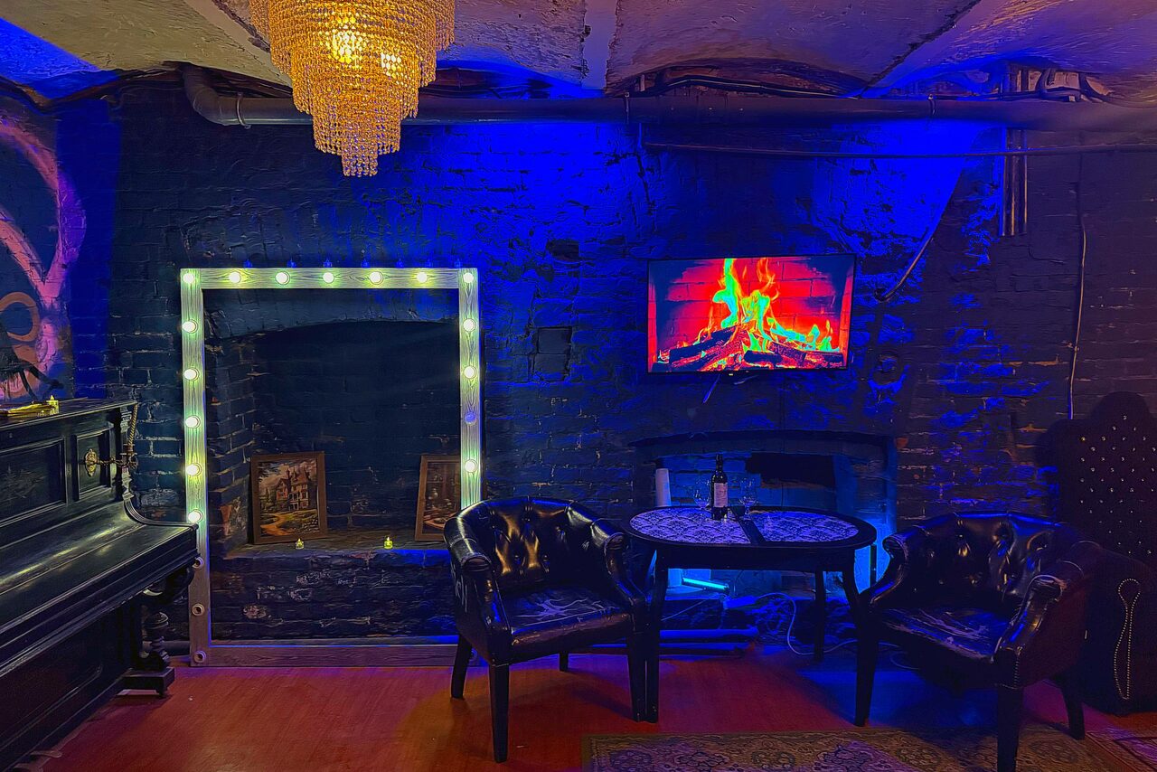 Арендовать Лофт «Рич» (RICH). Полуподвальное помещение с темными стенами, деревянный пол. Две кожаные кресла и круглый стол с бутылкой и бокалом. Синее освещение, крупное зеркало с лампами, декор в виде экрана с изображением камина, чёрный пианино, люстра.