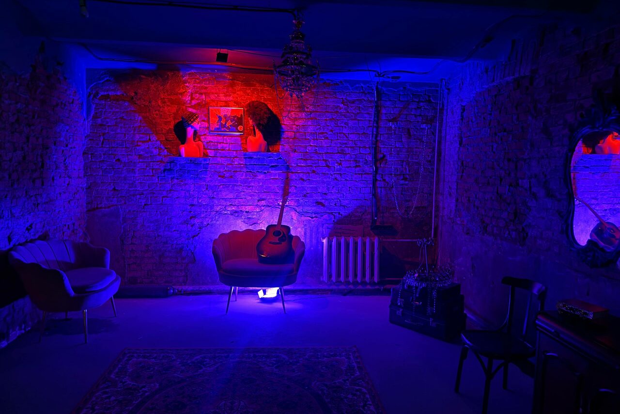 Арендовать Лофт «Рич» (RICH). Лофт с кирпичными стенами, фиолетовым освещением. Небольшой диван, кресло с гитарой, стул. Ковёр на полу, люстра, зеркало на стене. Радиатор вдоль стены, рисунок или картина. Текстуры из кирпича, дерево, текстиль.