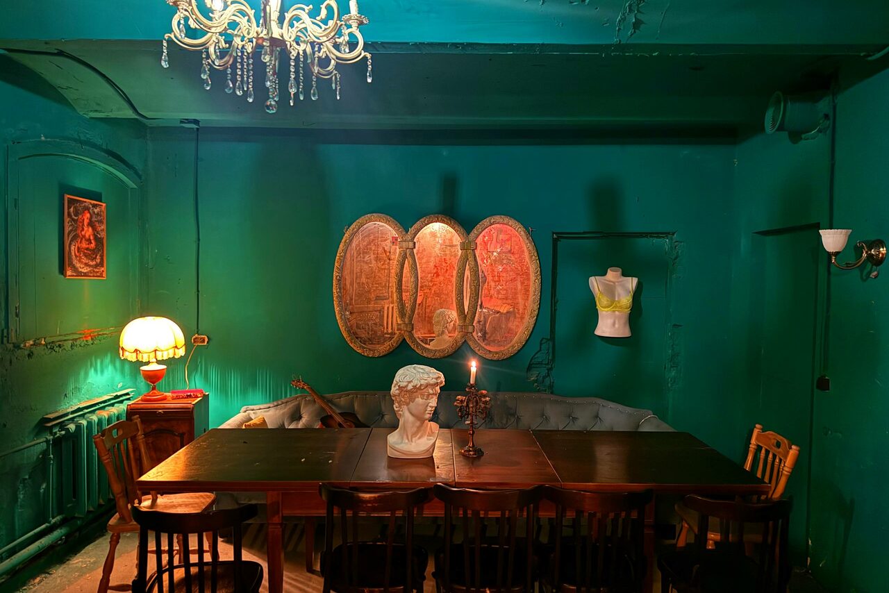 Арендовать Лофт «Рич» (RICH). Комната с зеленым интерьером, деревянный стол с восемью стульями, бюст и свеча на столе, люстра и настольная лампа, овальные картины на стене, манекен, зеркало. Потолок и стены зелёные, декоративные элементы, мягкое освещение.