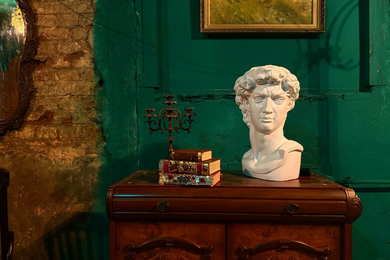 Арендовать Лофт «Рич» (RICH). Старинное помещение с кирпичной стеной и зелёной краской. На деревянной тумбе бюст, стопка книг и канделябр. Рама зеркала видна сбоку. Верхняя часть светло-зелёной стены с картиной в золотистой раме. Создано тёплое освещение.