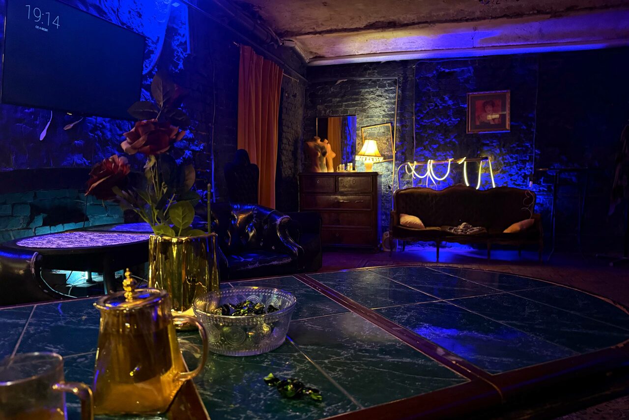 Арендовать Лофт «Рич» (RICH). Полутёмное помещение с диванами, круглым столом и зелёной мраморной столешницей. Чёрные кирпичные стены освещены синим светом. На столе золотистый чайник и стеклянная миска. Рядом кресло, лампа, зеркало и картина на стене.