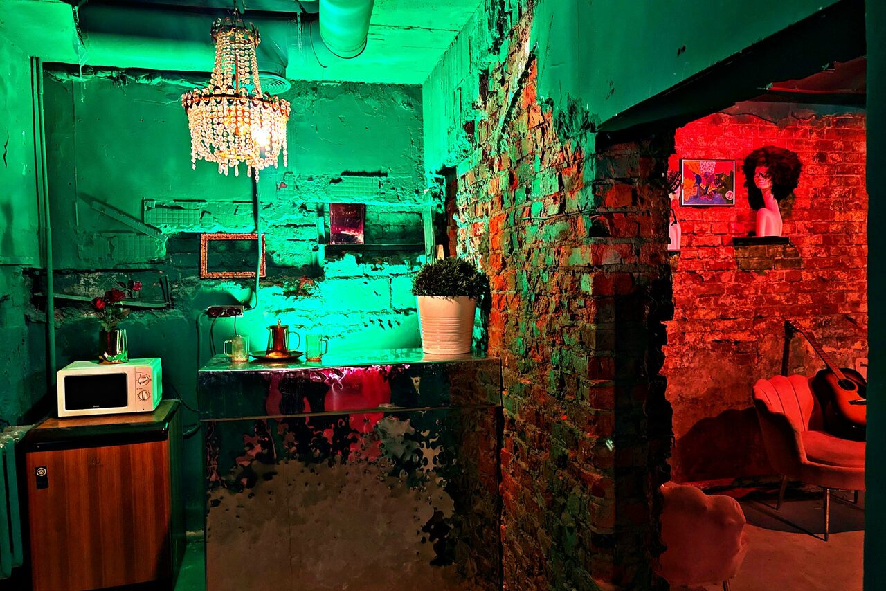 Арендовать Лофт «Рич» (RICH). Грубая кирпичная стена, барная стойка с зеркальной поверхностью, яркое разноцветное освещение, хрустальная люстра, микроволновая печь, растение в горшке, постеры на стене, в углу акустическая гитара.