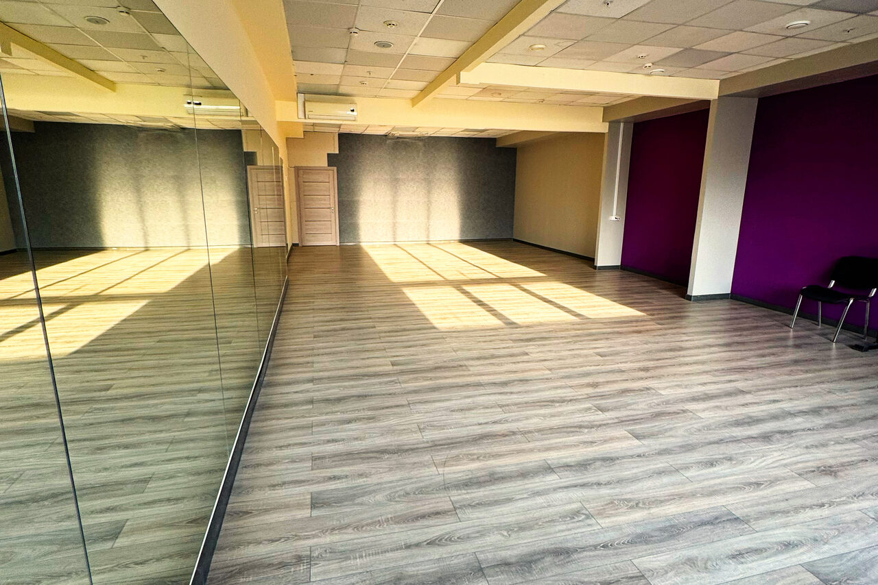 Арендовать Зал №10 «Сеть» 120 кв.м.. Просторное помещение с деревянным полом, зеркальной стеной слева, светлыми потолочными панелями с освещением. Одна стена окрашена в фиолетовый цвет, вдоль неё стоят два стула. Свет от больших окон падает на пол.