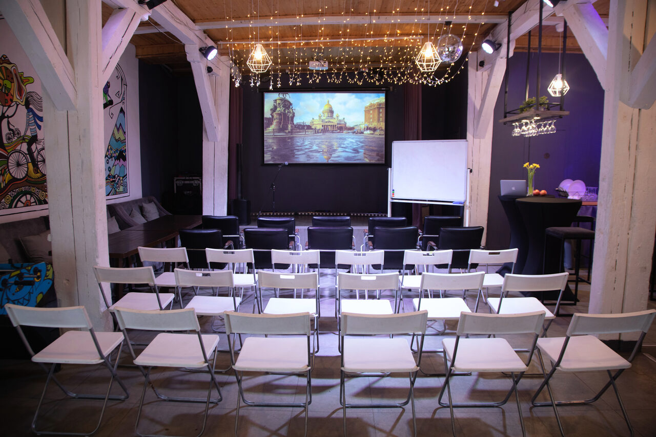 Арендовать Сатурн Лофт. Зал с деревянными балками и видом столов расположенными в форме аудитории. Экран для проекции, флипчарт, подвесные светильники, светодиодные гирлянды. Черные и белые стулья. Слева арт-шаблон на стене. Темно-серые и светло-серые тона интерьера.