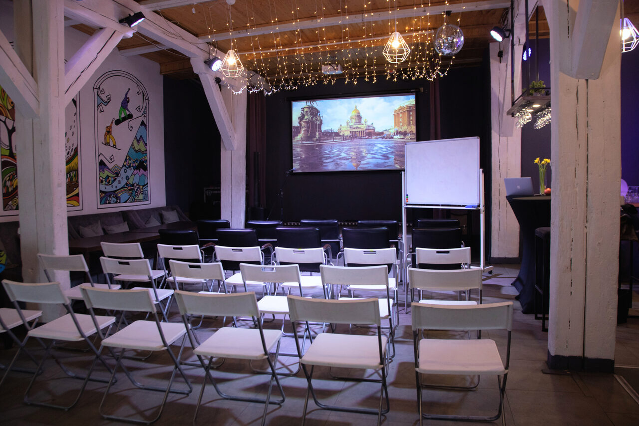 Арендовать Сатурн Лофт. Пространство с креслами, расположенными рядами перед экраном с изображением. Справа флипчарт, стойка с барными стульями. Деревянные балки и потолок. Настенные картины, гирлянды и подвесные лампы. Светлая и темная отделка, серые и белые тона.