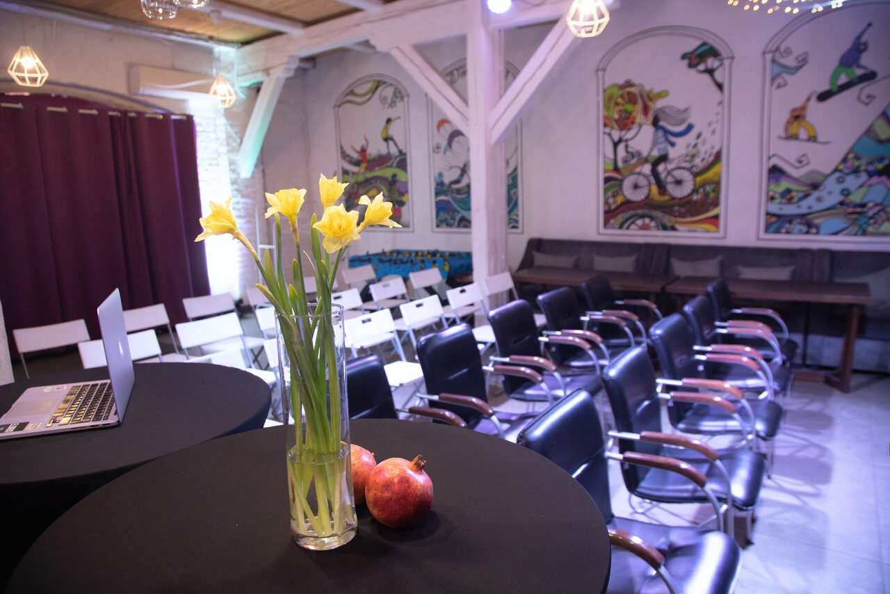 Арендовать Сатурн Лофт. Конференц-зал с креслами в нескольких рядах, столы с черными скатертями, ноутбук, ваза с желтыми цветами, гранаты, на стенах яркие настенные рисунки, фиолетовые занавески, мягкое освещение, балки на потолке.