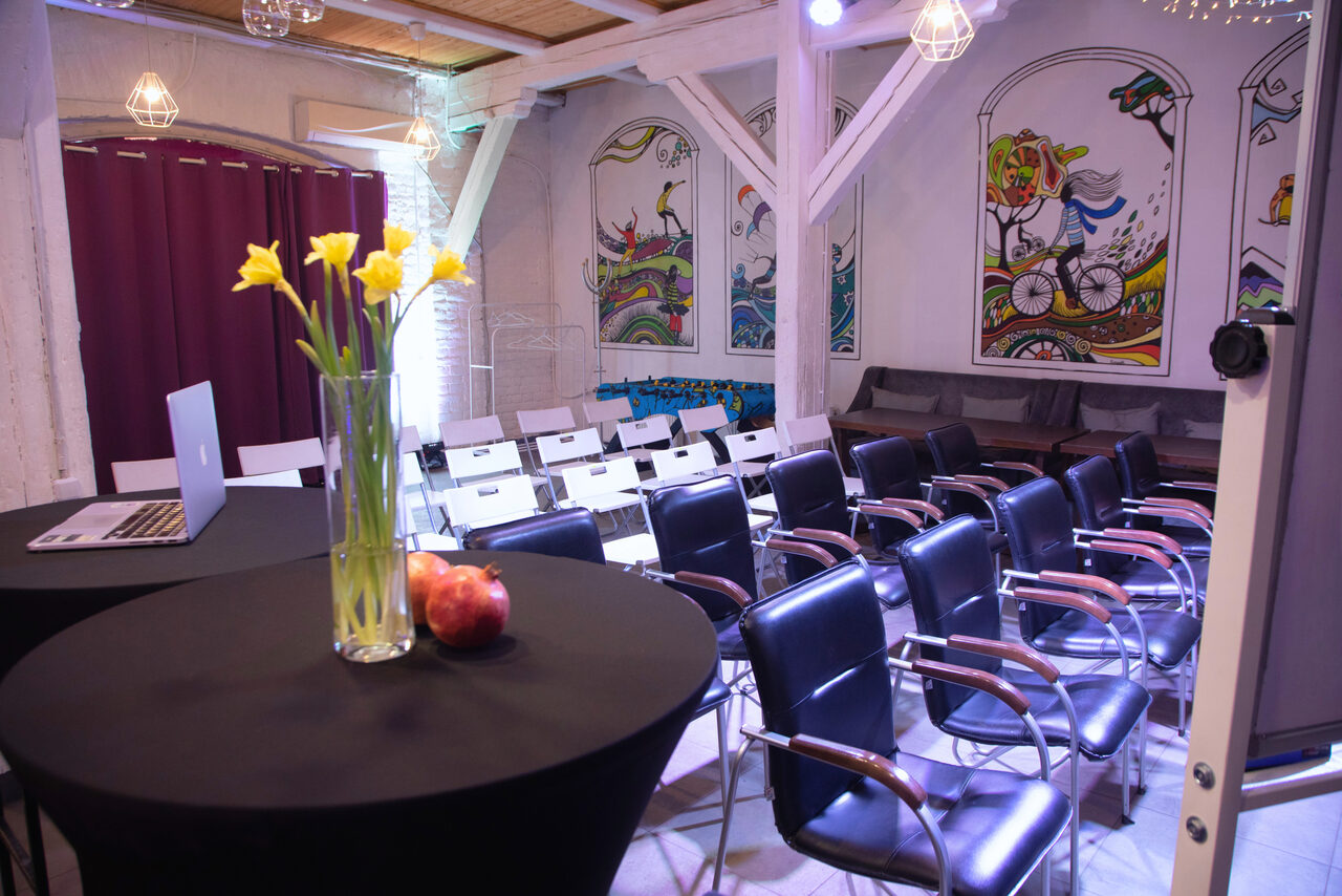 Арендовать Сатурн Лофт. Конференц-зал с рядами черных и белых стульев с деревянными подлокотниками. Стены украшены яркими настенными рисунками, потолок с деревянными панелями. В помещении высокий стол с черной скатертью, вазой с цветами и ноутбуком. Мягкое искусственное освещение.