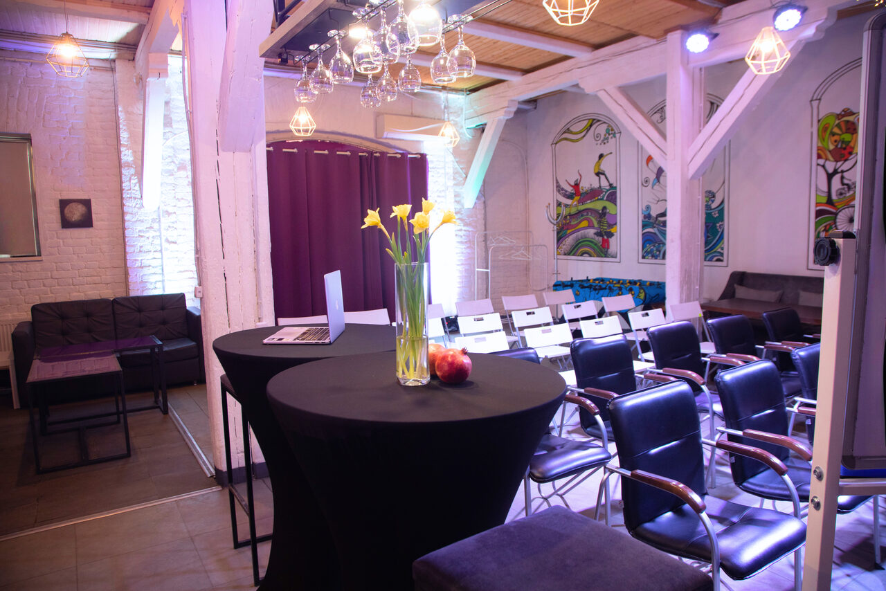 Арендовать Сатурн Лофт. Просторный зал с рядами черно-белых стульев и мягкой мебелью. Высокие круглые столы с вазой цветов и яблоками. Декоративные элементы на стенах, фонари подвешены под деревянным потолком. Белые кирпичные стены. Теплое и фиолетовое освещение.