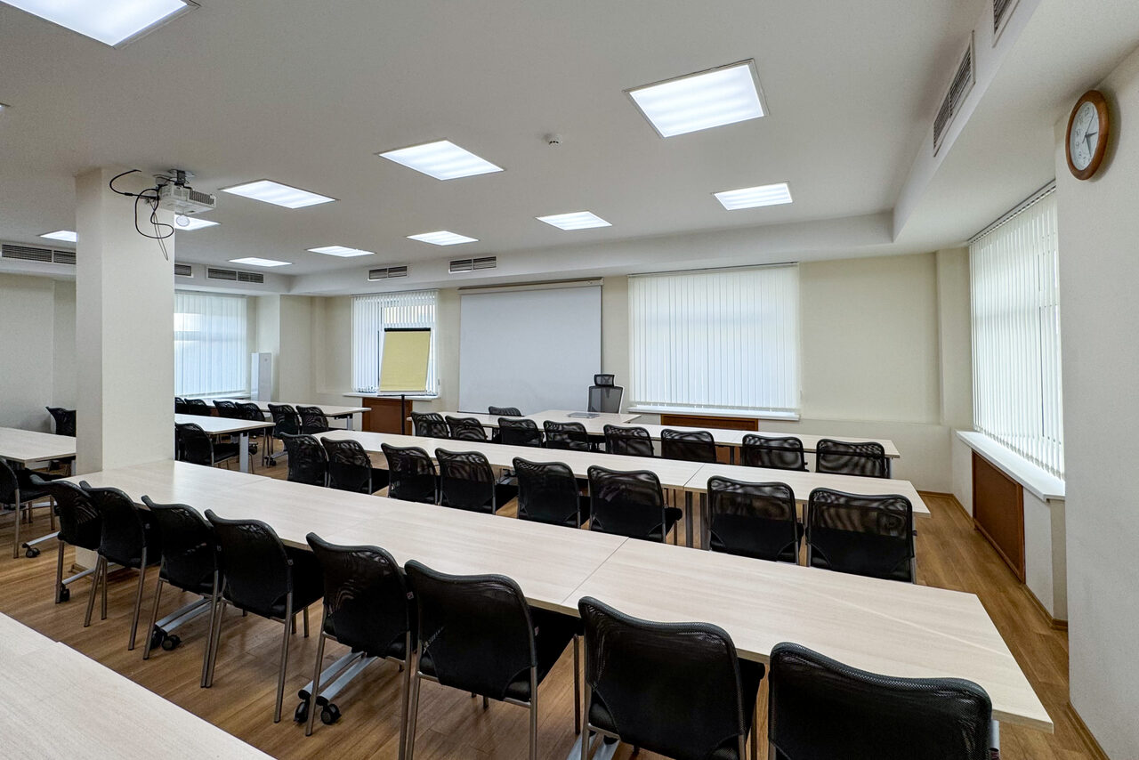 Арендовать Новое образование. Учебная аудитория. Аудиторное пространство с длинными светлыми столами и черными стульями на колесиках в три ряда. Белый экран, проектор и флипчарт. Потолочные светильники, окно с жалюзи, деревянный пол. Светло-бежевые стены, часы на стене.
