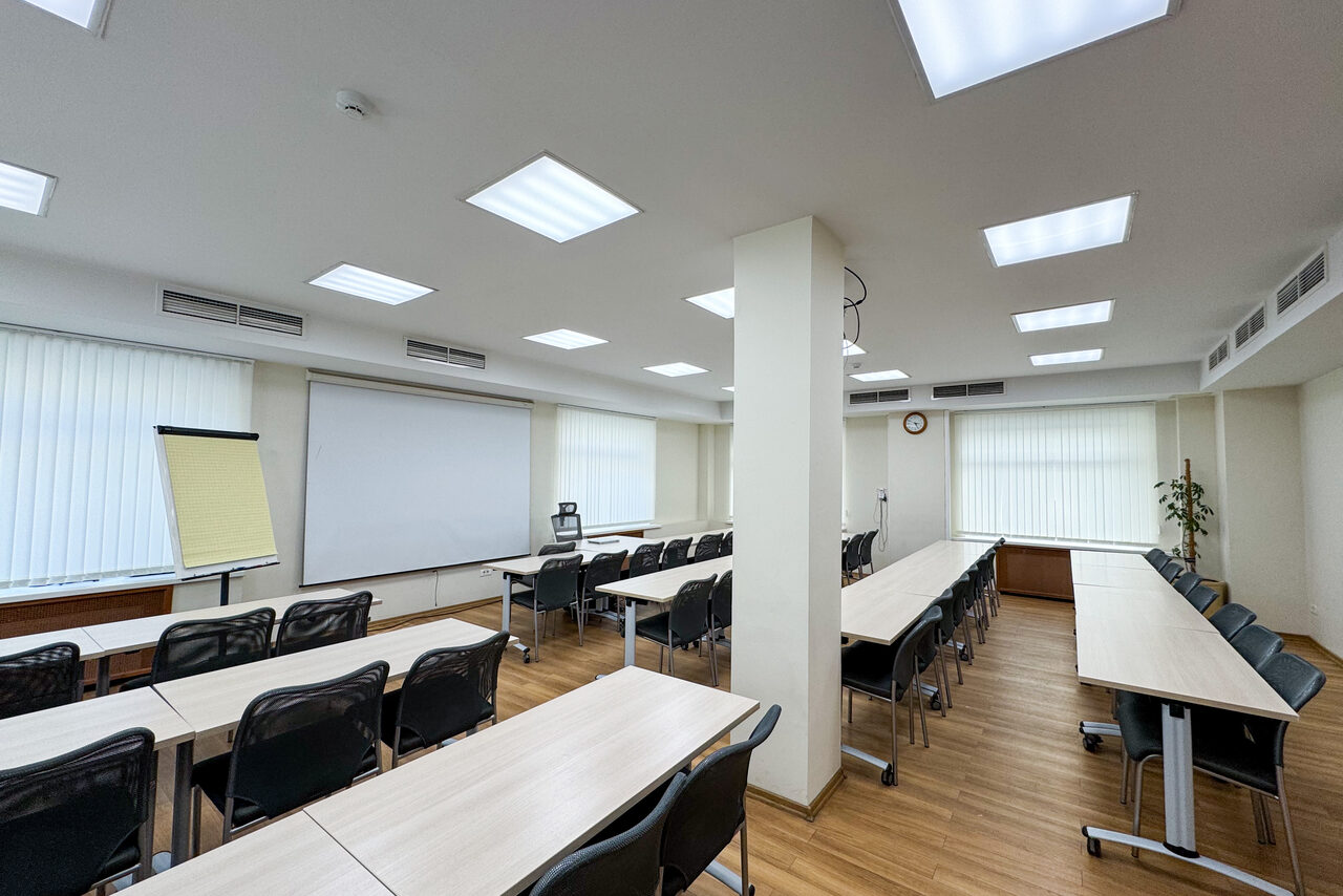Арендовать Новое образование. Учебная аудитория. Учебное пространство с рядами столов и стульев, проекционный экран, флипчарт и растение. Деревянные полы, светлые стены, потолочные светильники. Вентиляционные решетки и часы на стене. Панорамные окна с вертикальными жалюзи.