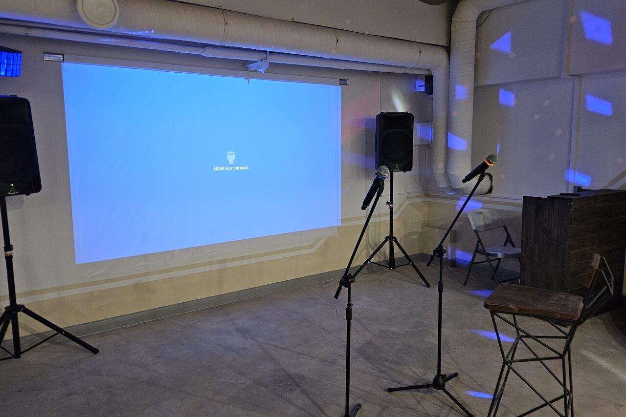 Арендовать Караоке-лофт с бильярдом. Помещение с бетонным полом и светлыми стенами. Проектор проецирует синий экран на стене. Два стула и стойки с микрофонами. Две колонки на стойках. Видны вентиляционные трубы на потолке. Светодиодные светильники создают узоры на стенах.