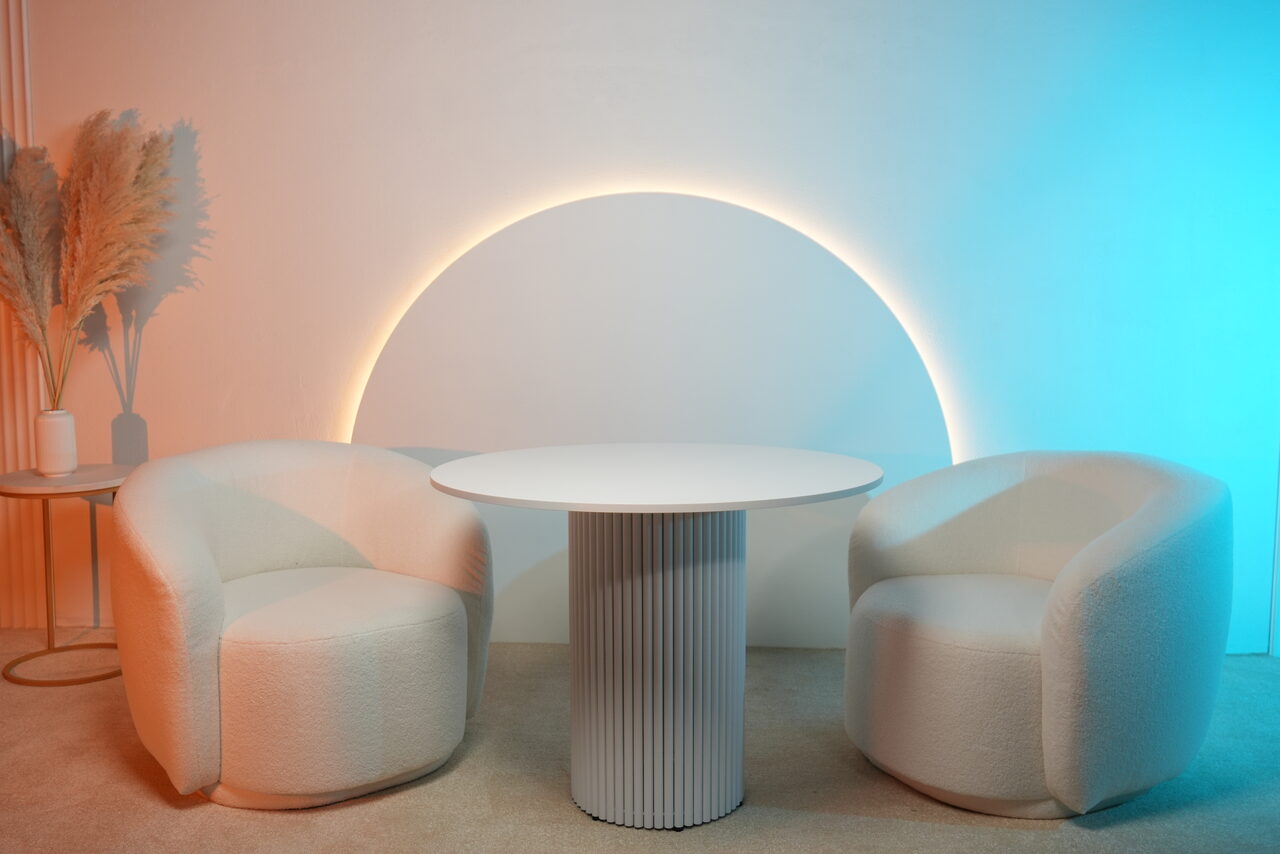 Арендовать Лофт Восторг. Современное пространство с двумя мягкими креслами и круглым столом с рифленой ножкой. Стена с подсветкой в виде полукруга, цветное освещение, декор в виде сухоцветов на круглом столике. Дизайн выполнен в светлых тонах.