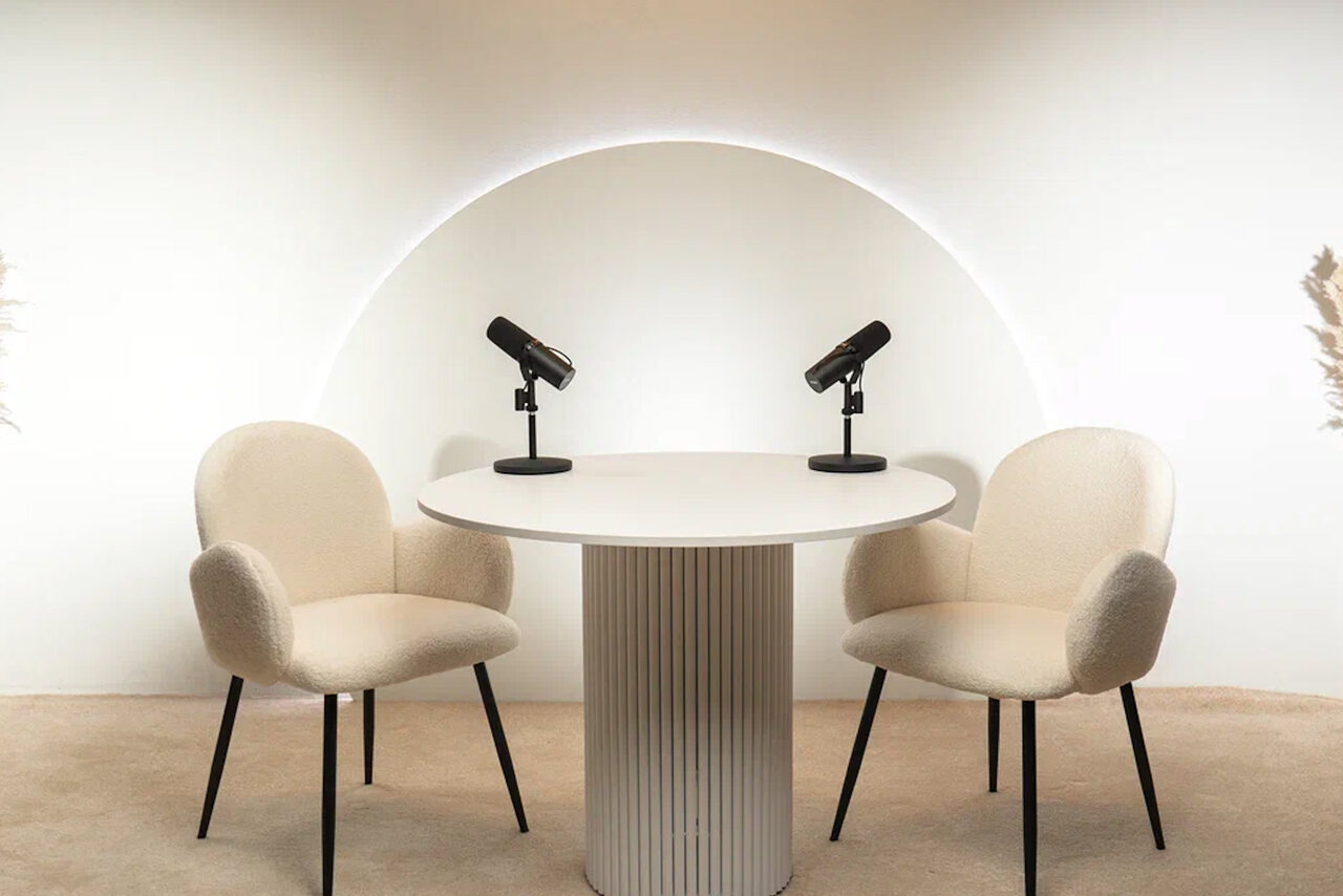 Арендовать Лофт Восторг. Круглый стол с двумя креслами светлого цвета перед белой стеной с круглой подсветкой. На столе установлены два микрофона. Напольное покрытие бежевого цвета. С обеих сторон от стола находятся декорации из сухих растений на полу.