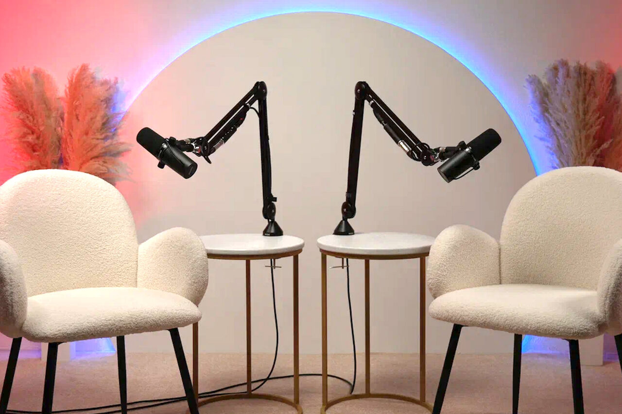 Арендовать Лофт Восторг. Две белые кресла с округлыми подлокотниками расположены напротив круглых столов с черными микрофонами на стойках. Фон подсвечен красным и синим. По бокам стоят декоративные растения. Стены светлые.