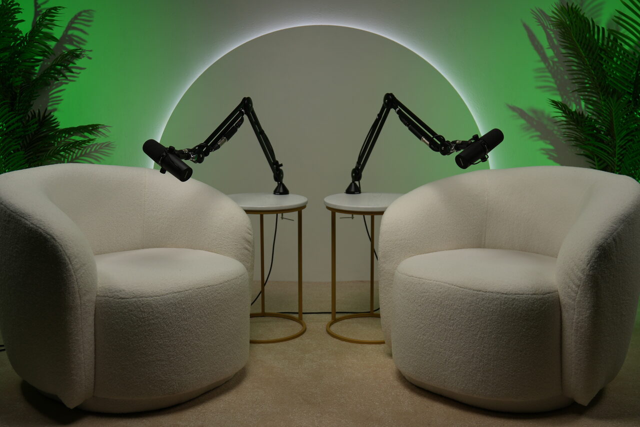 Арендовать Лофт Восторг. Две белые кресла-коконы перед двумя круглыми столиками с черными микрофонными стойками; задний фон с полукруглым светодиодным освещением на стене, зелёные растения по краям, пол бежевый.