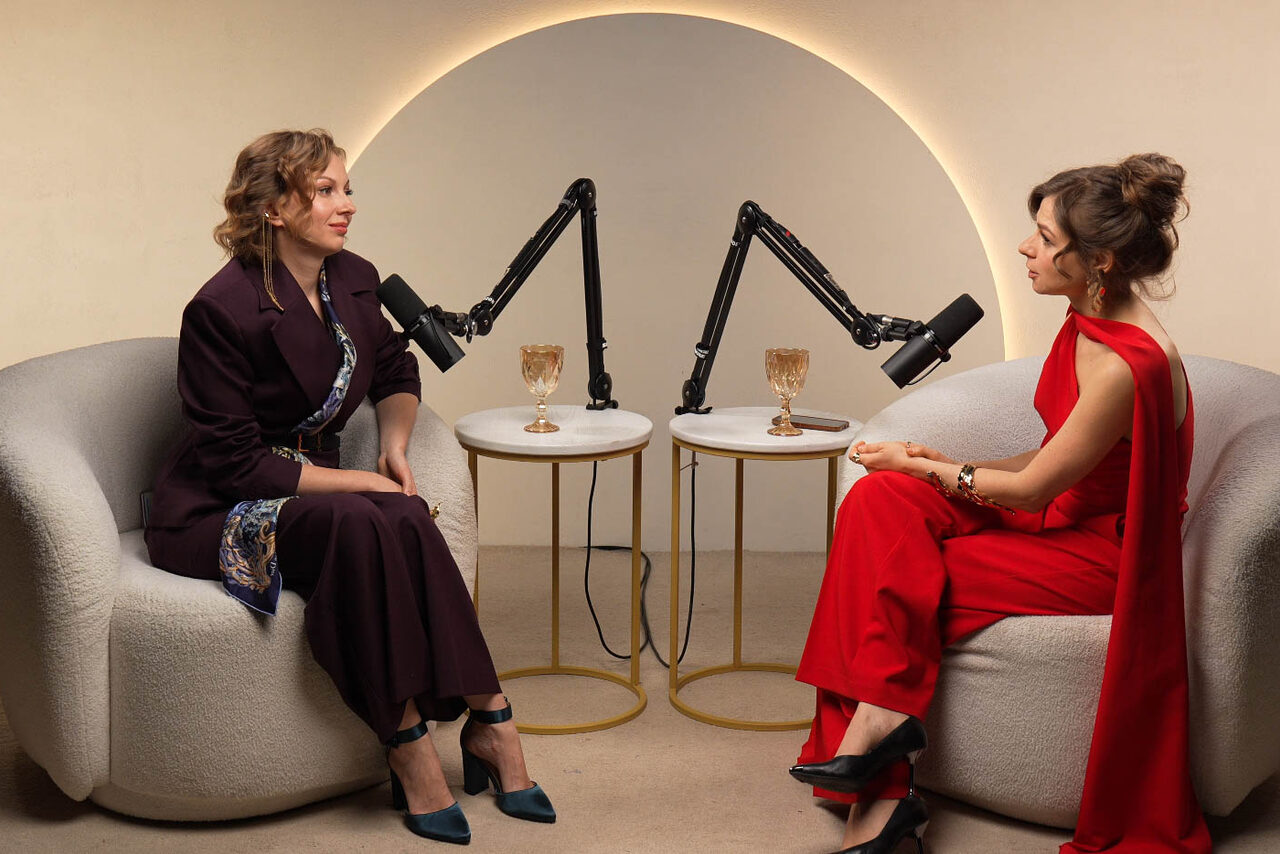 Арендовать Лофт Восторг. Современное помещение для интервью с двумя креслами светло-серого цвета, двумя круглыми столами с золотыми ножками, микрофонами на стойках и подсветкой на стене. На столах стоят бокалы. Освещение теплое, стены нейтральных тонов.