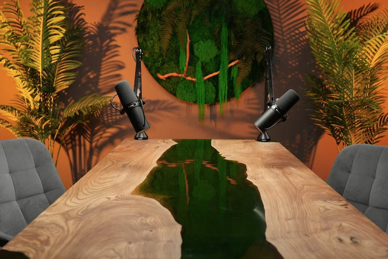 Арендовать Лофт Восторг. Современное помещение с прямоугольным деревянным столом с эпоксидной вставкой, двумя микрофонами на стойках, серыми стульями и зелеными растениями. На стене круглая инсталляция с мхом. Освещение теплое, стены оранжевые.