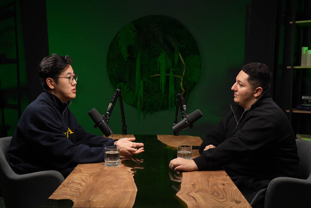 Арендовать Лофт Восторг. Студийное пространство с деревянным столом неправильной формы, двумя микрофонами на стойках. Два человека сидят напротив друг друга на серых креслах. Задний фон с круглым зелёным декоративным элементом. На столе стоят два стакана с водой.