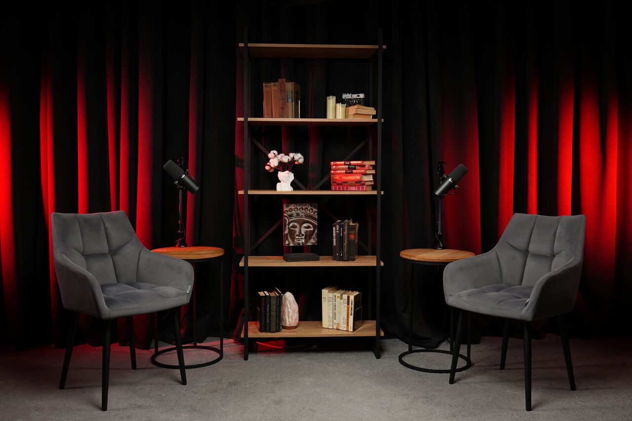 Арендовать Лофт Восторг. Пространство с двумя серыми креслами, стеллажом с книгами и декором, двумя круглыми столиками и микрофонами. Задний план — красные и черные шторы. Освещение теплое, контрастное. Пол серый, материал неясен.