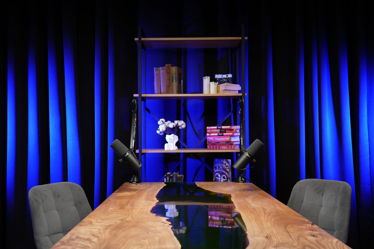 Арендовать Лофт Восторг. Маленькая комната с деревянным столом, двумя серыми стульями, микрофонами, стеллажом с книгами и украшениями. Задний план синими шторами. Освещение мягкое, направлено вверх, подчеркивает задний фон.